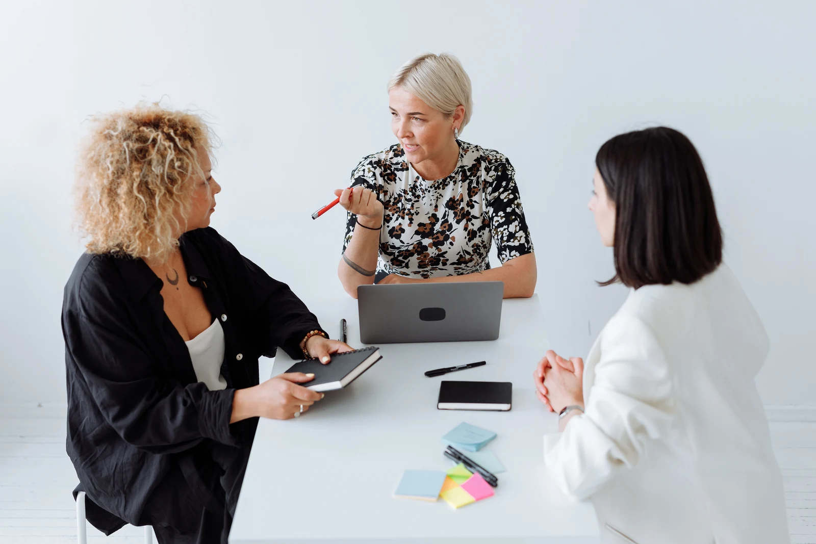 Three women in a bright office meeting around a laptop, one listening carefully while another speaks