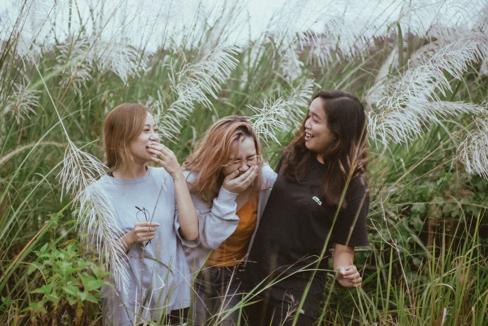 Three women laughing together outdoors in long grass, showing the value of community during perimenopause